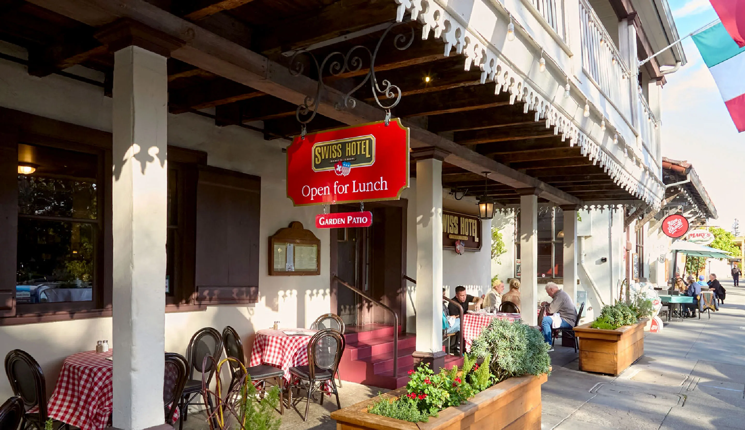 Swiss Hotel's shaded front porch with gingham-topped tables and an Open for Lunch sign, guests dining on the sidewalk