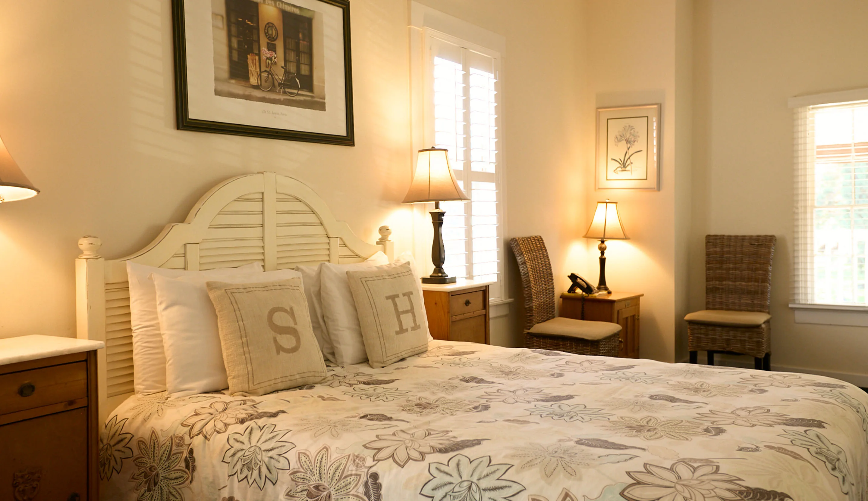Room 1 at the Swiss Hotel, with a queen bed in a floral quilt and S and H pillows, wicker chairs and soft lamplight on cream walls