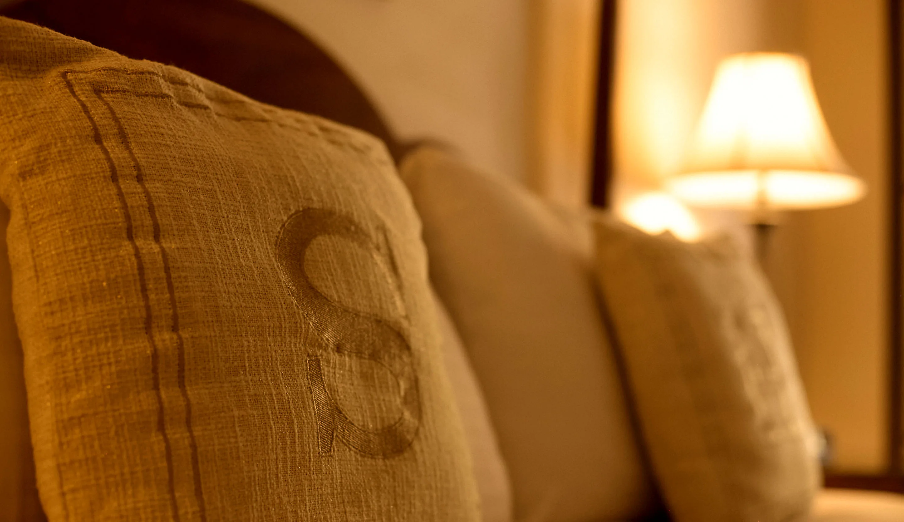 Close-up of a linen pillow embroidered with an S on the bed in Room 5 at the Swiss Hotel, lit by a warm bedside lamp